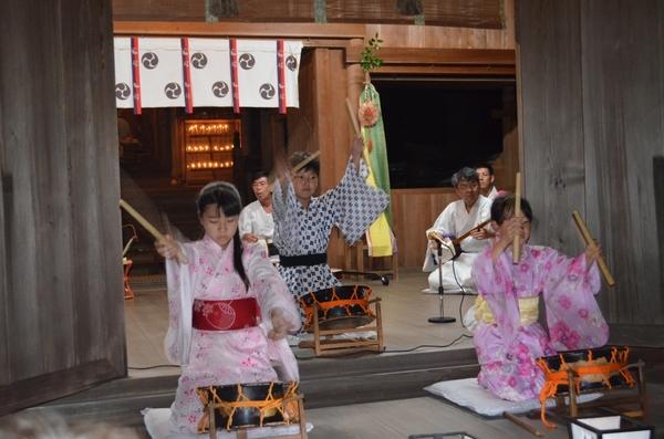 浴衣を着た女の子と男の子が締太鼓を叩いてその後ろで大人たちが三味線を弾ている写真